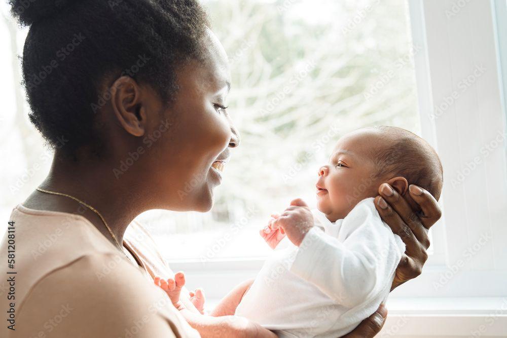 A mother holds her newborn close, standing in front of a window that lets in warm, natural light.