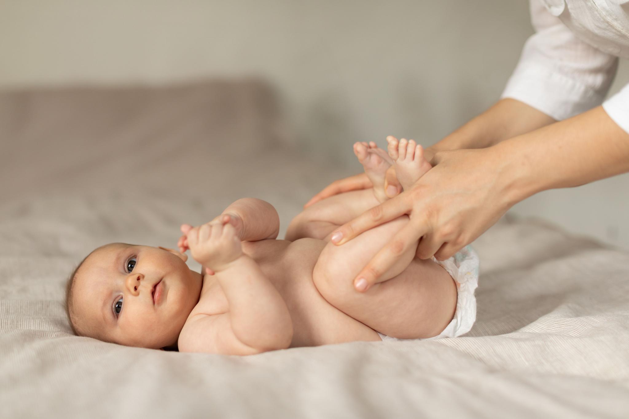 A newborn baby lying on its back on a light-colored blanket. An adult's hands, likely the mother's or caregiver's, are gently holding and massaging the baby's legs and feet. The baby is awake and looking towards the camera