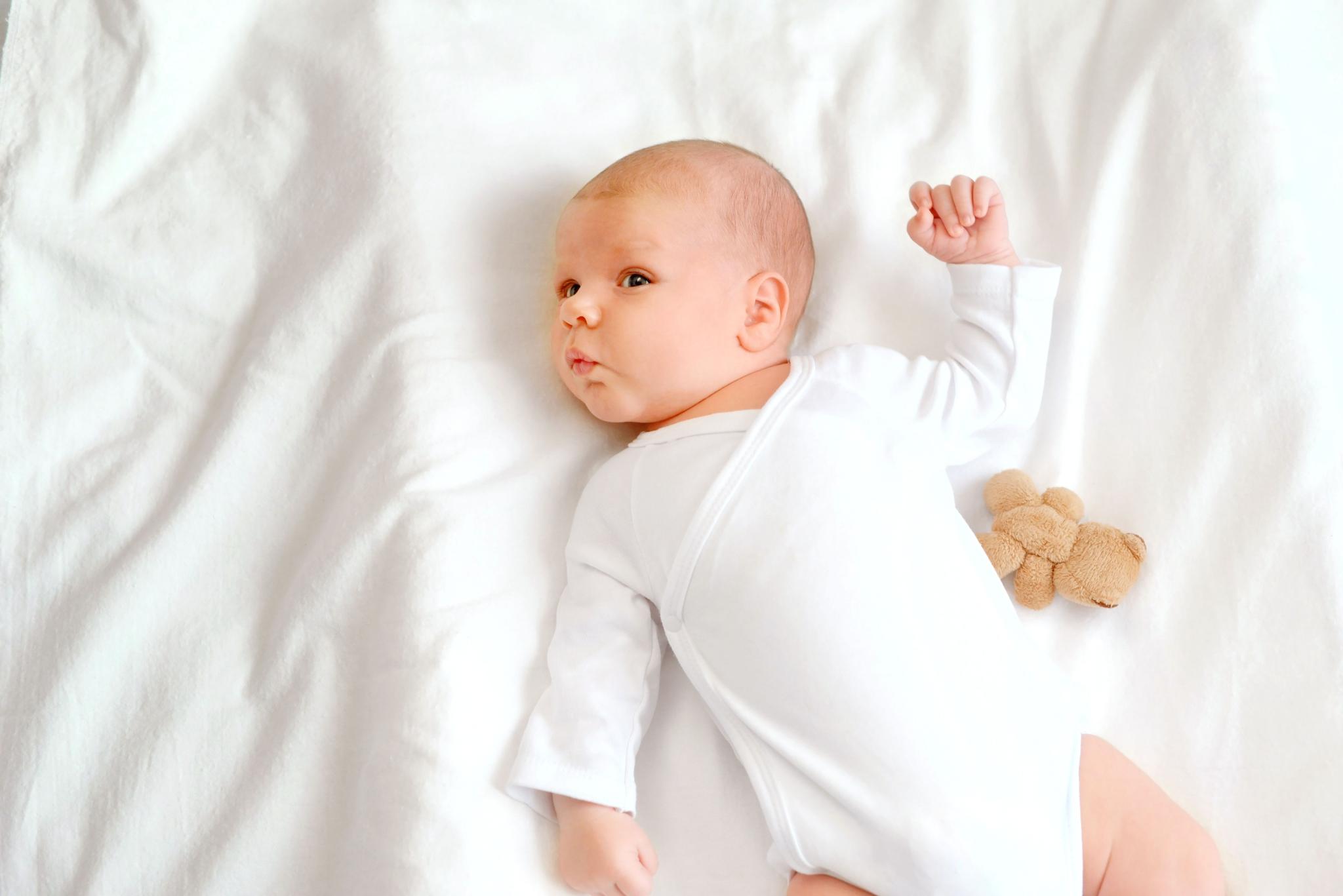 A baby lies on a bed, hands on its face, looking curious. Soft bedding surrounds the little one.